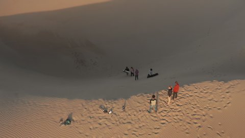 4K California nature landscape. Aerial slow motion video of a group of young people in the desert at sunset. Travelers in colorful sports wear are sliding from the top of golden sand dune, Oceano
