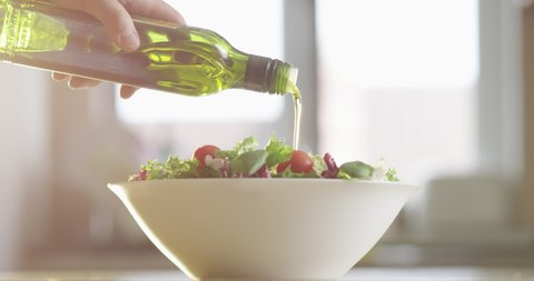 Olive Oil is Pouring into a Plate of Vegetable Salad with Lettuce and Cherry Tomatoes on a Kitchen Table with Sun Flares
