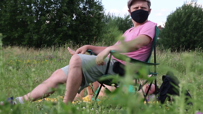 A man in a protective mask and cap sits in a camping chair. A woman is sunbathing on the ground. Outdoor recreation in self-isolation during a pandemic.