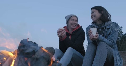 Two Cute Girls Sitting on Log on Seashore. Before them is Burning Bonfire. Bright Flame. Girls Drink Hot Tea from White Cups. Couple. Cute to each other. Smiling. Look at each other. In love. Camping