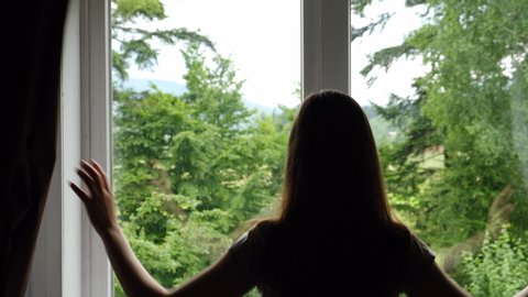 Rear view of silhouette young woman opening curtain lace looking through window. Confident unrecognizable lady enjoying watching beautiful green forest view, enjoys an active summer weekend at home