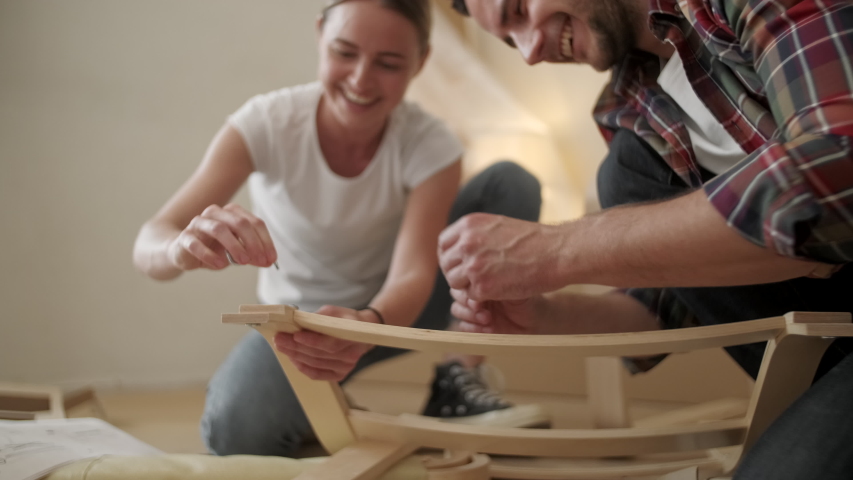 Portrait Happy Smiling Couple Collects Furniture as a Team. Girl Helps to Assemble Chair Details. Female Tightens Bolt. Moving to New Apartment, Young Family Assemble Furniture Royalty-Free Stock Footage #1055389865