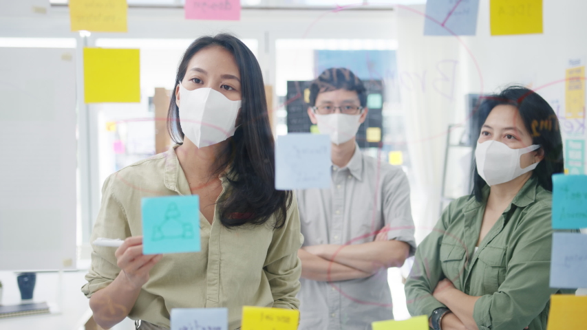 Asia young businesspeople discussing business brainstorming meeting working together sharing data and writing glass wall with medical face mask back at work in office. Life and work after coronavirus. | Shutterstock HD Video #1055397734