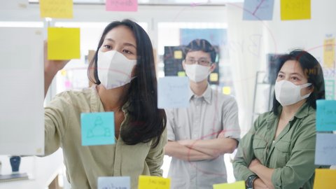 Asia young businesspeople discussing business brainstorming meeting working together sharing data and writing glass wall with medical face mask back at work in office. Life and work after coronavirus.