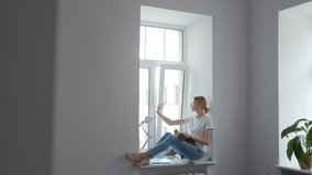 Girl sitting on windowsill in room with white walls with cat on her lap taking a picture or shoots video on her phone for social networks