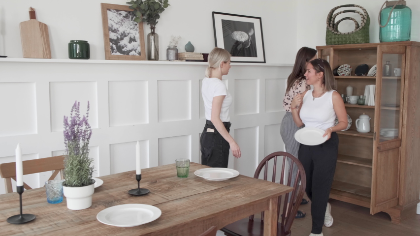 Young woman setting the table in living room putting water carafe for meal while happy friends talking. Happy people group enjoy modern home party dinner or lunch. Handheld video Royalty-Free Stock Footage #1055401571
