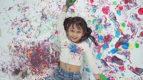 Top view of adorable smiling asian little girl is laying down and playing her painted hands on her abstract artwork at home, concept of freedom, free play and art education for kid learning at home.