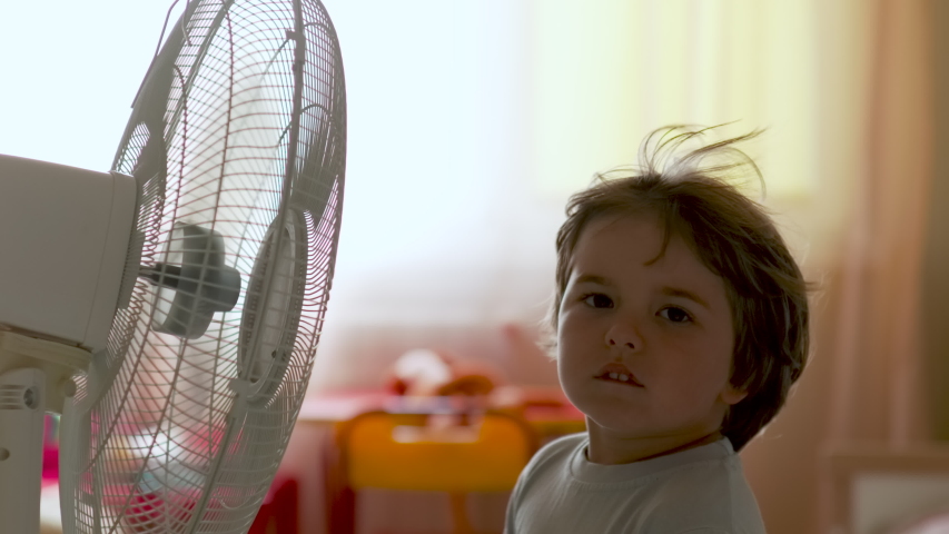 Boy Standing in Front of Fan. Child Enjoying Cool Wind From Electric Fan at Home at Summer Vacation. Suffer From Heat High Temperature in Front of Ventilator Cooling Herself With Electric Fan-Cooler. Royalty-Free Stock Footage #1055404376
