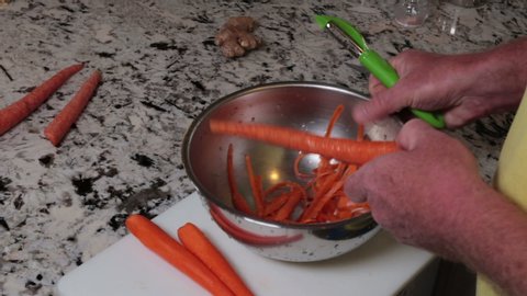 Male hands peeling organic carrots onto a white cutting board placed on a stone kitchen counter. Human hands removing the outer layer of a carrot with a peeling utensil preparing food for a recipe.