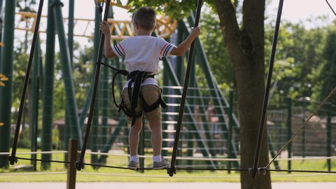 A brave little boy moves along the ropes between the trees, uses a safety rope