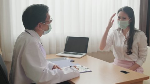 Asian female patient coming to examination room and consult health problem to male doctor sitting in room. Medic man, woman wearing protective surgical face mask preventing covid virus pandemic.