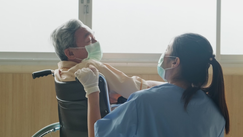 Asian nurse taking care of mature male patient sitting on wheelchair in hospital. Young woman and old man wearing surgical face mask for protection of covid 19 pandemic. Girl smile to elderly man. Royalty-Free Stock Footage #1055407649