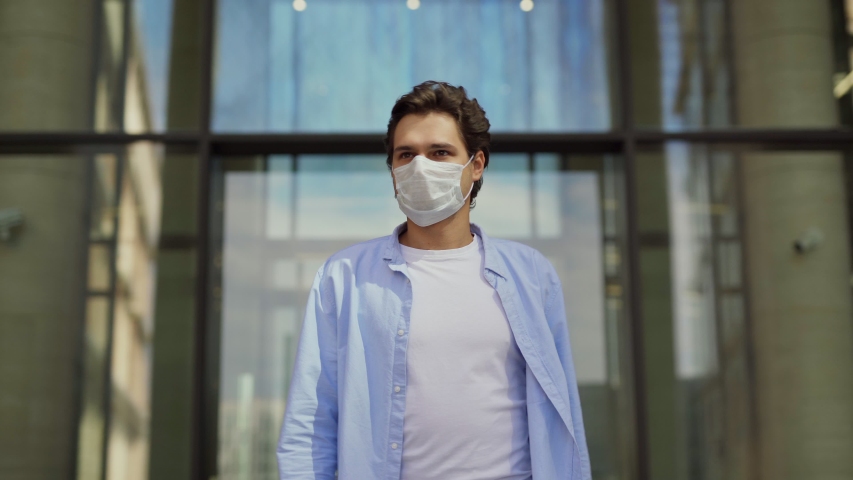 Front view of young man leaving office building and taking off medical face mask outdoors