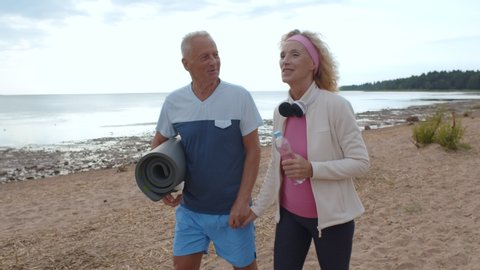 Стоковое видео: Senior sporty couple holding hands walking on empty beach relaxing after workout. Aged man and woman strolling on shore carrying fitness mat and discussing exercising outdoors
