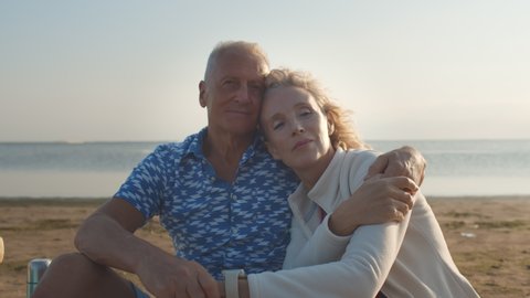 Loving senior couple hugging sitting on sea beach outdoor. Happy elderly man and woman enjoying retirement and life. Concept of wellbeing, happiness, male and female health, tenderness.