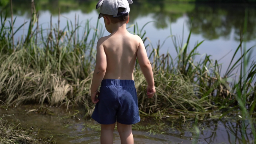 A small boy, a European, dressed in blue shorts and a cap stands in the river with his back to the camera, turns, takes a stick from the Bank and puts it in the water