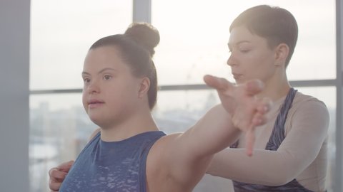 Cheerful young disabled woman with down syndrome doing yoga exercise with her personal trainer helping her