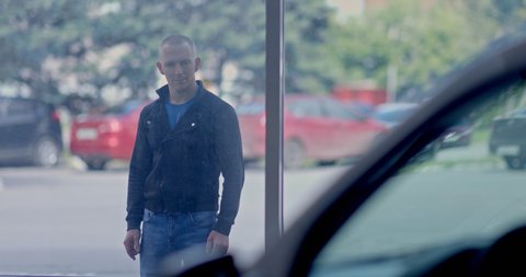 Young man looks at the car through the showcase of a car dealership. Buying a car, a man chooses a car. Portrait view.