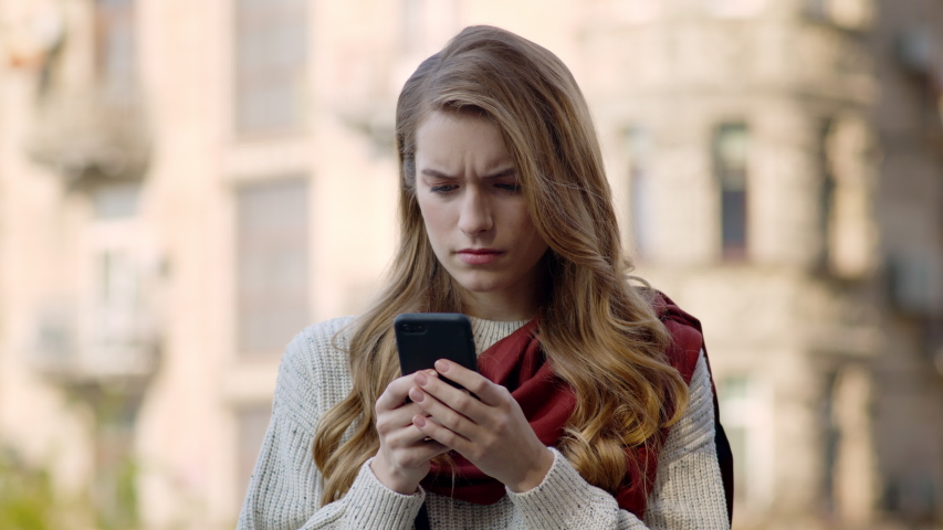 Portrait of shocked woman holding mobile phone in hands outdoors. Closeup upset hipster girl looking smartphone screen on city street. Disappointed female person using cellphone in urban background. Royalty-Free Stock Footage #1055421296