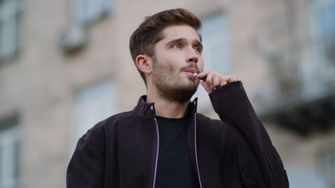 Portrait of handsome man smoking electronic cigarette in urban background. Close up thoughtful hipster man vaping on city street. Relaxed guy using e-cigarette outdoors.