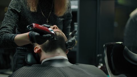 Woman hairdresser works with trimmer with a beard of a client. Close-up of young man's face with not thick beard. Unrecognizable barber girl in black gloves professionally makes a haircut.