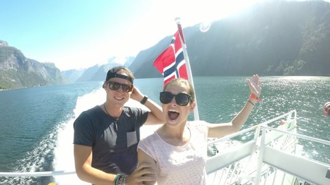 Couple on a fjord boat tour with a Norwegian flag waving behind them, Songefjorden Norway. The motor of the ship makes the water wavy and foamy. Lush green mountains surrounding the fjord. Strong wind