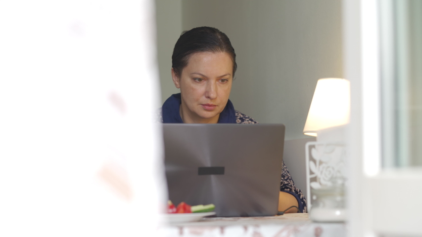 Confident adult woman working with laptop from home in the morning, woman in dressing gown having breakfast using notebook in kitchen, distant job online working from home.