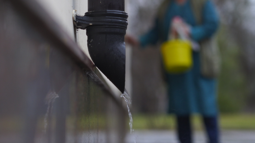 Rain water flowing from rain drain pipe on a building wall close-up, silhouette of a housewife woman in background. Royalty-Free Stock Footage #1055426795