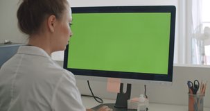 Young woman doctor work on computer with green mockup screen. Girl in uniform and lab coat clothes sitting in hospital office room using laptop. Over shoulder camera shot. 4k video footage slow motion