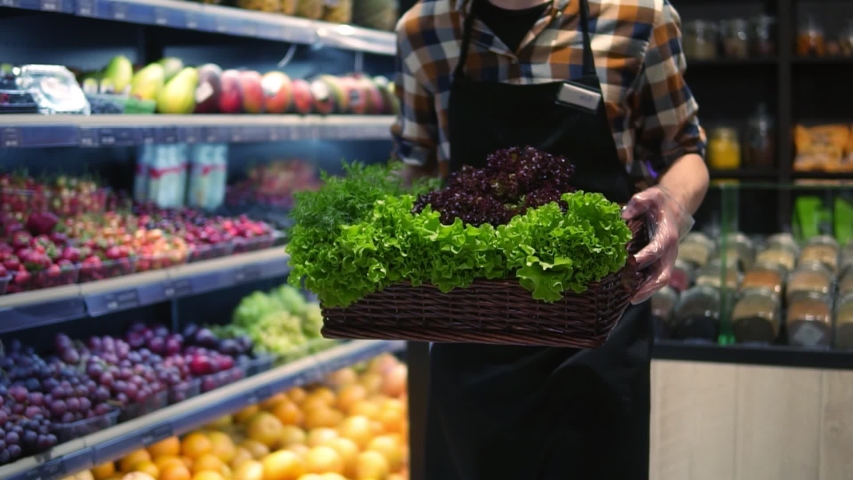 Seller man in apron in supermarket walking by vegetables aisle with box of fresh greens to arrange. Caucasian worker in local supermarket holding box of greens. Close up. Slow motion Royalty-Free Stock Footage #1055430056