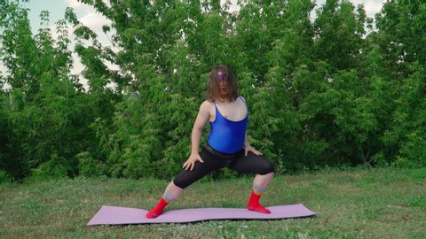 Funny Fat Man with Long Hair in a Tight Female T-shirt Is Engaged in Martial Arts on fitness mat on Green Lawn in City Park. Playful Guy Overweight Depicting a Girl. Sport humor concept.