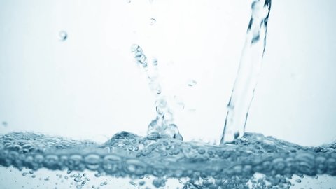 Water stream in slow motion falls on smooth clean surface, creating air bubbles, drop splashes after falling. Freshness of a clear blue liquid poured into transparent container on white background