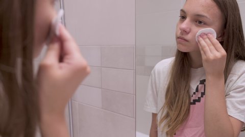 Teen girl looks at her face in bathroom mirror and using cotton pads. Beautiful teenager with problem skin. Health care on facial concept.