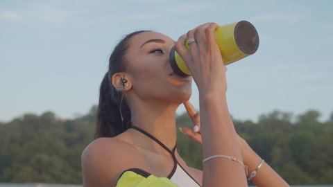 Lifestyle portrait of positive beautiful african american sportswoman drinking energy drink from thermos mug in morning glow after outdoor workout at sunrise, expressing wellness and happiness.