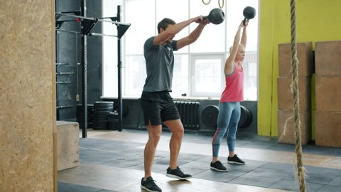 Slow motion of young couple guy and girl exercising in gym squatting with kettlebells working out together. Crossfit, active lifestyle and people concept.