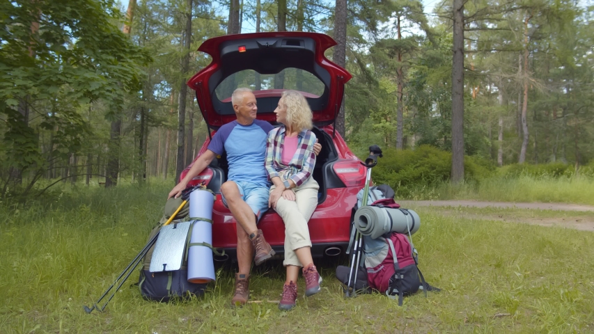 Portrait of mature man and woman hikers sitting in open car trunk and hugging in forest. Active aged couple traveling by car. Happy retirement, tourism lifestyle, love and travel concept