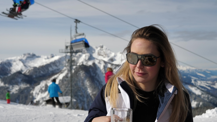 Medium closeup of a Caucasian woman with brown hair wearing sunglasses and drinking beer at a skiing resort with skiing cable cars and snow covered mountains in the background