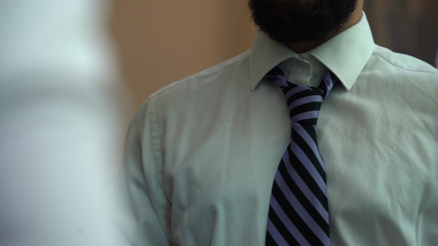 Man tying a tie. Businessman getting dressed. Man  in white shirt,  tying a tie, stands by the mirror. Man's face not visible. Groom before wedding ceremony. Working time. Royalty-Free Stock Footage #1055448266