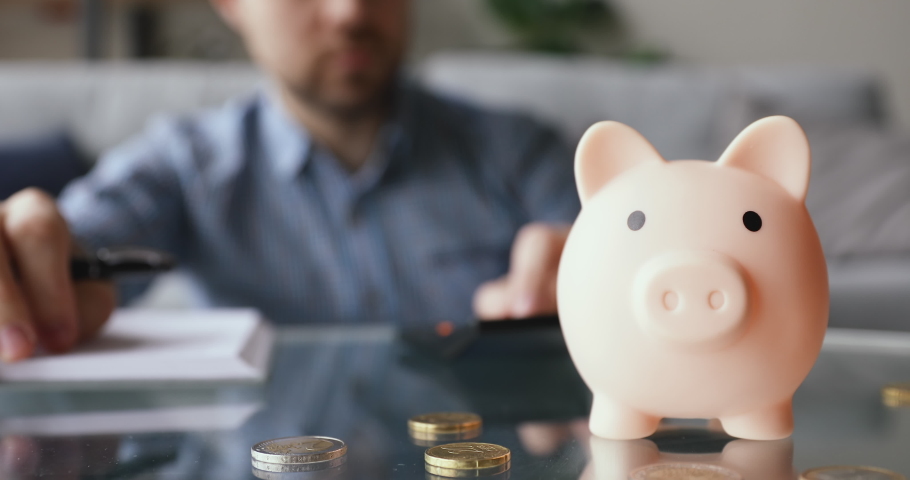 On background man calculates writing in daily planner personal expenses and incomes, close up focus on piggy bank euro coins on table, symbol of budget managing, make savings and take care of tomorrow | Shutterstock HD Video #1055448758