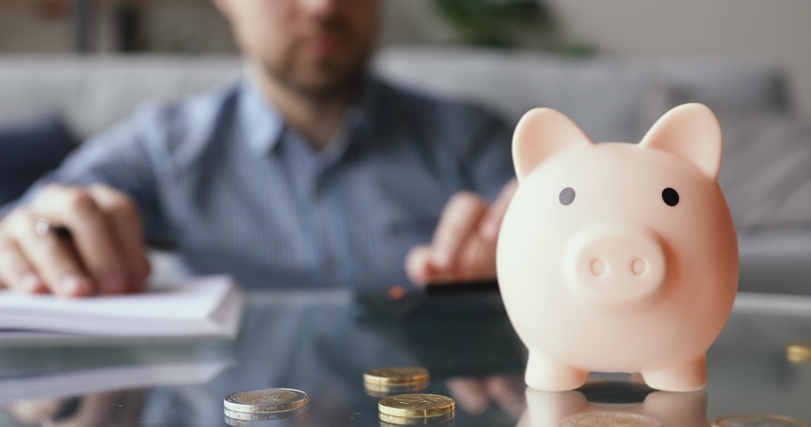 On background man calculates writing in daily planner personal expenses and incomes, close up focus on piggy bank euro coins on table, symbol of budget managing, make savings and take care of tomorrow | Shutterstock HD Video #1055448758