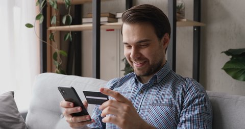 Smiling man sitting on couch holding cellphone using credit card makes instant payment feels happy. Easy on-line shopping, modern tech, safe successful convenient payment, sale and discounts concept