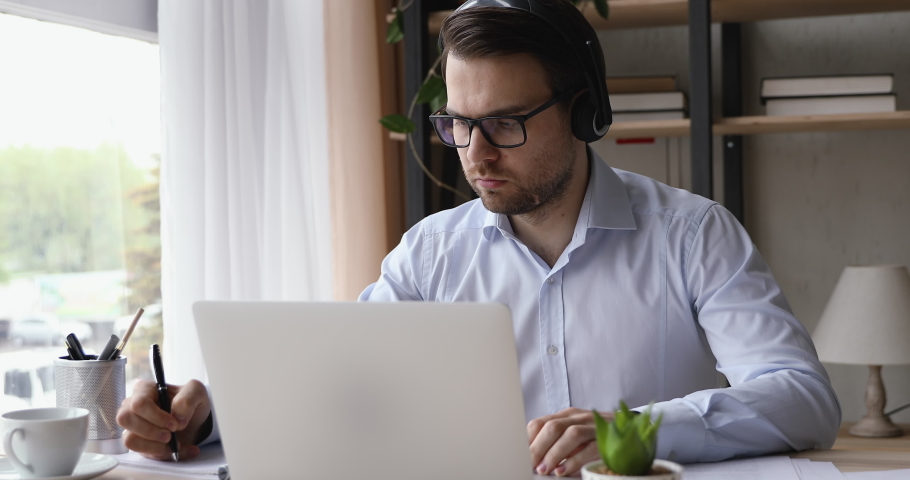 Serious man wear headset look ap pc screen writes ideas questions do review make research sit at desk. Businessman learn language improve knowledge, self-education, modern tech educational app concept Royalty-Free Stock Footage #1055448764