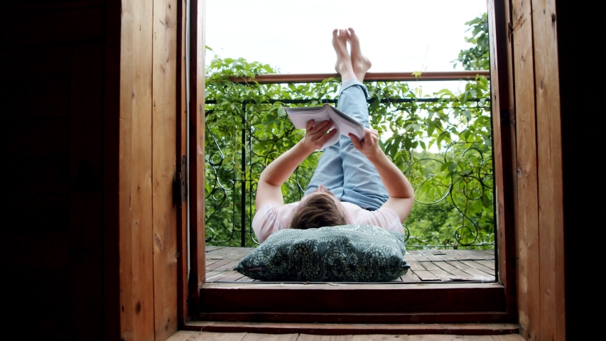 a man lies on his balcony and reads a book with his legs up. social distance during the coronavirus pandemic. time to get smarter Royalty-Free Stock Footage #1055451110