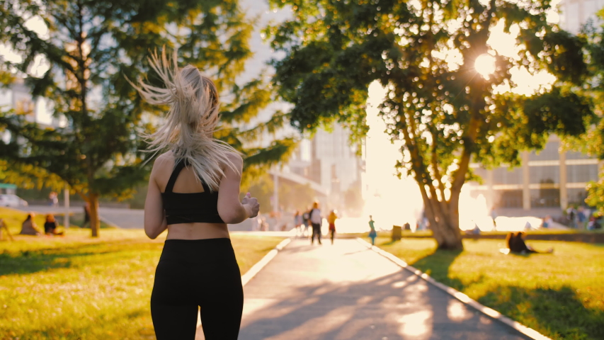 Slim young blonde woman in black sports bra and leggings jogging in summer park. Sporty sexy woman running, exercising outdoors at sunrise. Sun lens flare glare, healthy active lifestyle, back view
