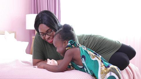 African American child with her mother saving money in piggy bank in bedroom. Happy african girl sitting with her mother inserting coins In pink piggy bank at home.