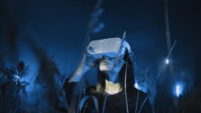 Girl uses VR glasses or headset at modern interactive exhibition in blue neon light outdoor, among wheat field. Future technology, 3D graphics, virtual and augmented reality, concept