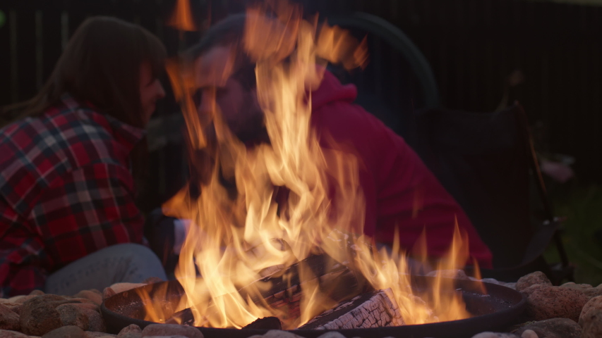 Young inlove couple sits near a fire after sunset and kiss. Husband and wife on a picnic in the evening by the fire. Happy leisure on weekend. Fire closeup macro shot. Soft focus.