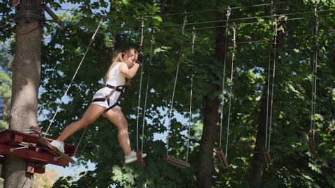 Young teen brunette overcomes obstacles in the rope town. A girl moves at a height along the ropes stretched between the trees, uses safety rope