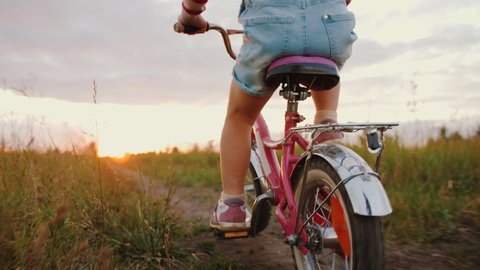 Little girl riding bike in field at sunset. Kid dressed in denim overalls rides bicycle on meadow trail, to pedal close up having fun outdoors. Family leisure, active holidays, childhood.