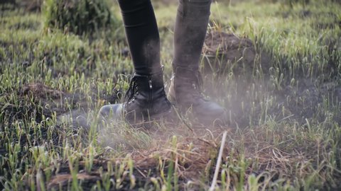 Human legs in stylish leather shoes spin in a circle on ashen burnt grass in slow motion in sunny weather. Person is spinning on the burned earth in a field during the day, smoke blowing after a fire.